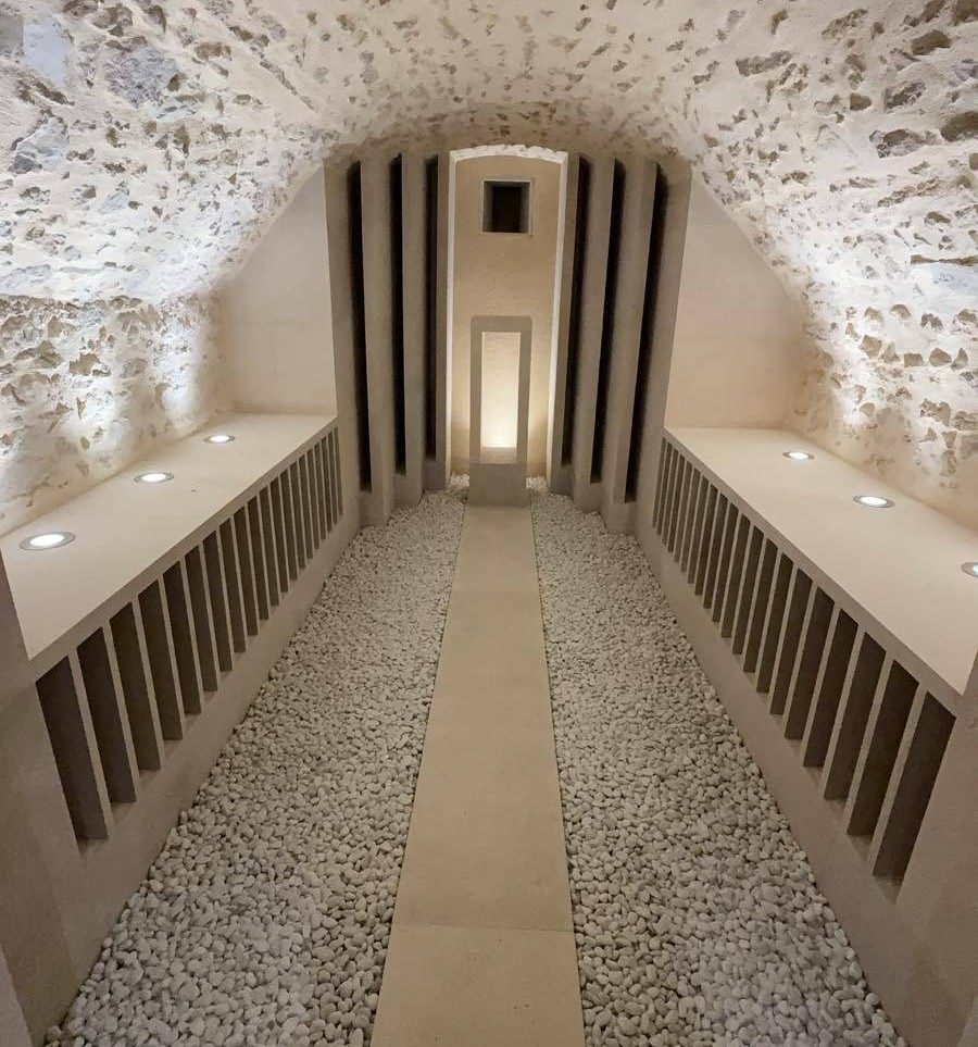 Vue en perspective d'une cave à vin voûtée entièrement rénovée, alliant l'ancien et le moderne. Le plafond en pierres apparentes rétroéclairé surplombe un aménagement contemporain composé de casiers à bouteilles verticaux épurés, d'un sol en gravier blanc avec une allée centrale en dalles de pierre, et d'un éclairage doux intégré.