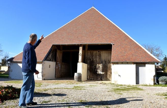 Un homme de dos pointe du doigt le sommet d'une grande grange traditionnelle dont la toiture en forme de pyramide vient d'être rénovée. Le toit est recouvert de tuiles plates neuves dans les tons ocre et brun. Le bâtiment présente des murs blancs en pierre, de larges portes en bois ancien et se situe sous un ciel bleu dégagé.